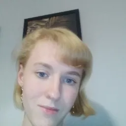 Portret młodej kobiety z blond włosami i delikatnym makijażem, pozującej w pomieszczeniu z obrazem w czarnej ramie na ścianie.