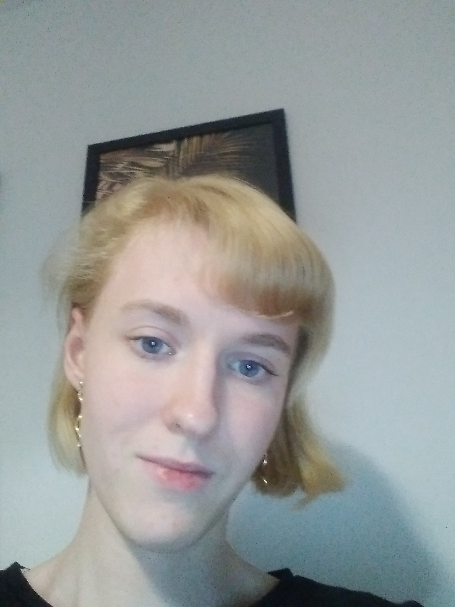 Portret młodej kobiety z blond włosami i delikatnym makijażem, pozującej w pomieszczeniu z obrazem w czarnej ramie na ścianie.