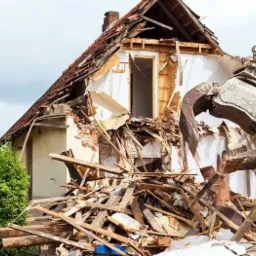 Żółty sprzęt ciężki z metalowymi szczękami chwyta elementy rozbieranego domu z ceglanym kominem i częściowo zniszczonym dachem z czerwonej dachówki. W tle zielona roślinność.