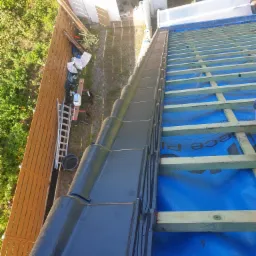 Układanie czarnej dachówki ceramicznej na łatach pokrytych niebieską membraną dachową, widok z góry na ogrodzenie z desek i budynek gospodarczy w tle.