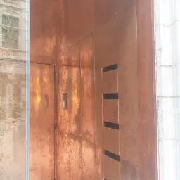 Miedziane wykończenie elewacji budynku z drzwiami i panelem z otworami wentylacyjnymi, widok z bliska z odbiciem budynku w szklanej powierzchni.