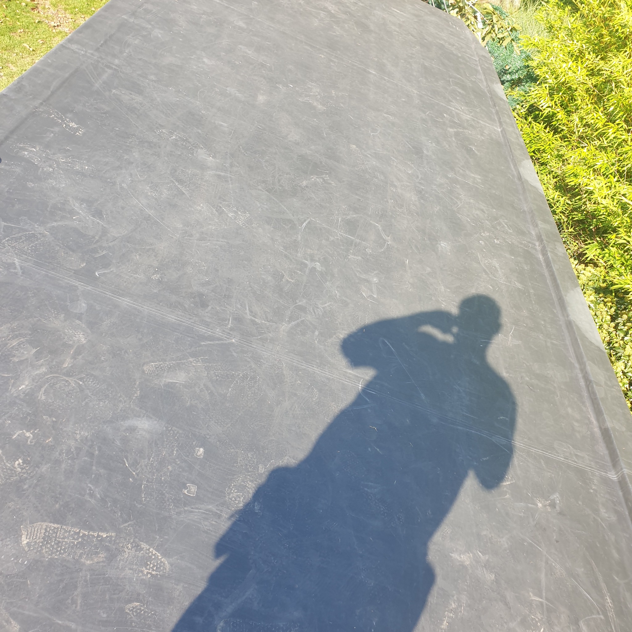 Szara, lekko zabrudzona powierzchnia dachu z widocznym cieniem osoby robiącej zdjęcie, obok bujna zieleń.