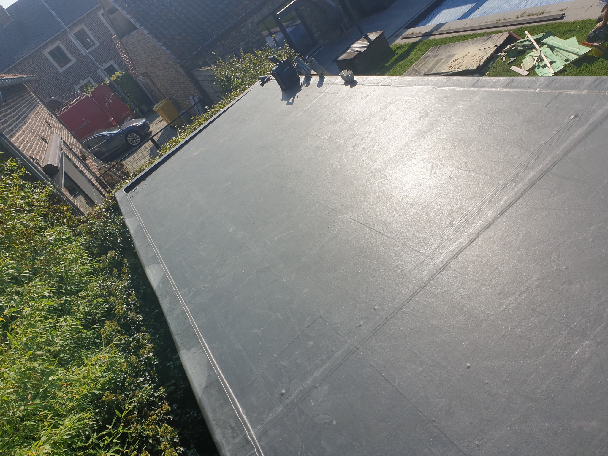 Płaski, szary dach pokryty papą termozgrzewalną z widocznymi zgrzewami, narzędzia dekarskie i resztki materiałów na krawędzi, zarośla i budynki w tle.