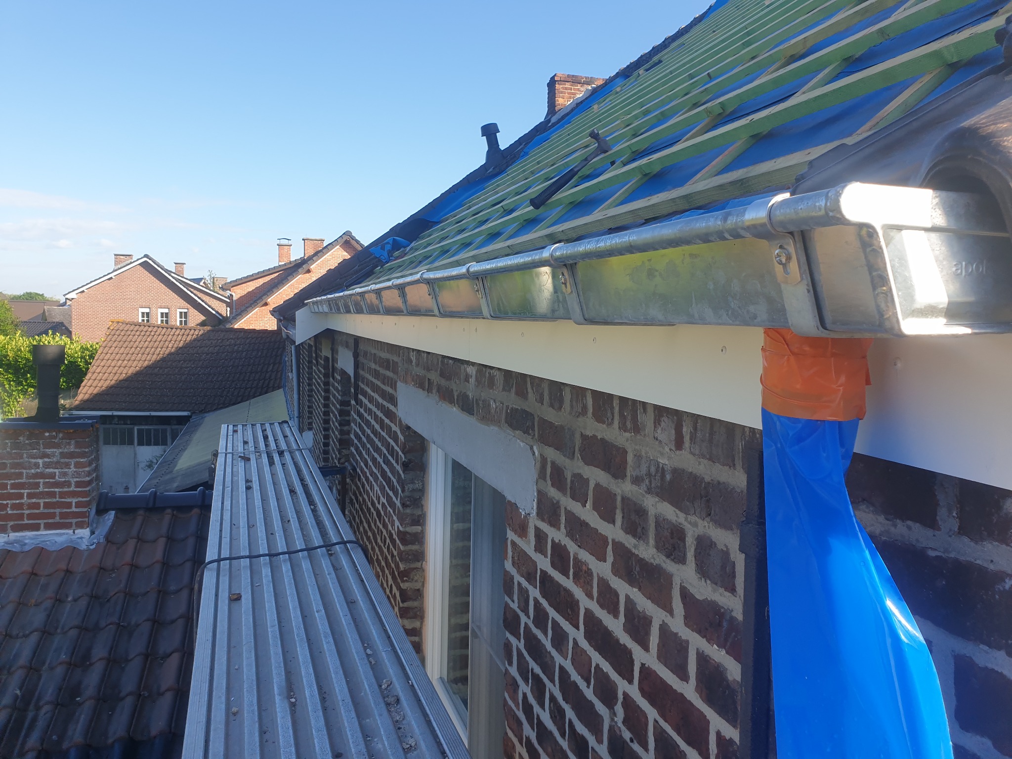 Montaż metalowej rynny na tle ceglanej ściany domu, widoczne rusztowanie i elementy konstrukcji dachu z zielonymi łatami i niebieską folią ochronną.