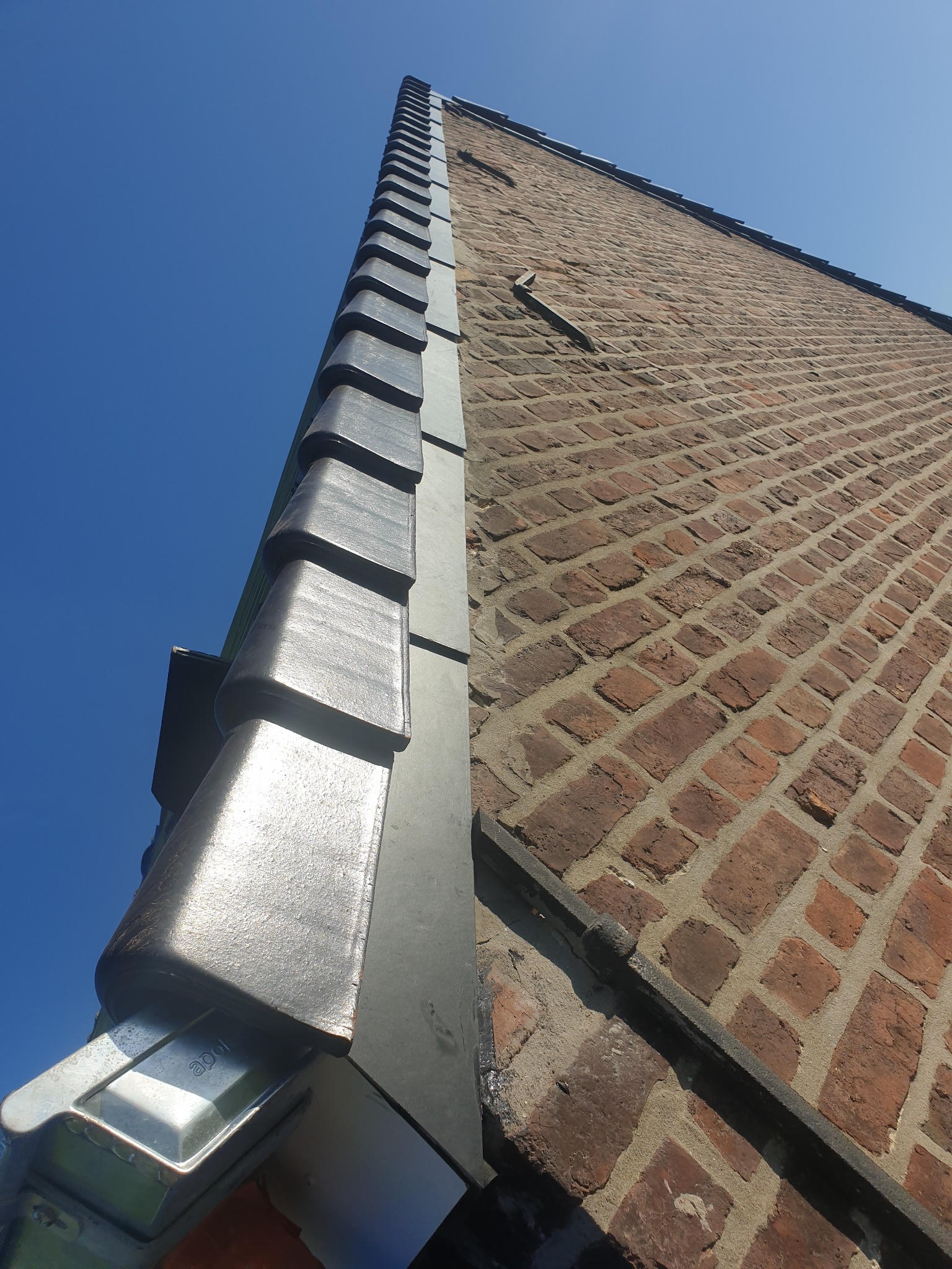 Widok z dołu na fragment muru z czerwonej cegły oraz metalowe obramowanie dachu z rynnami na tle błękitnego nieba.