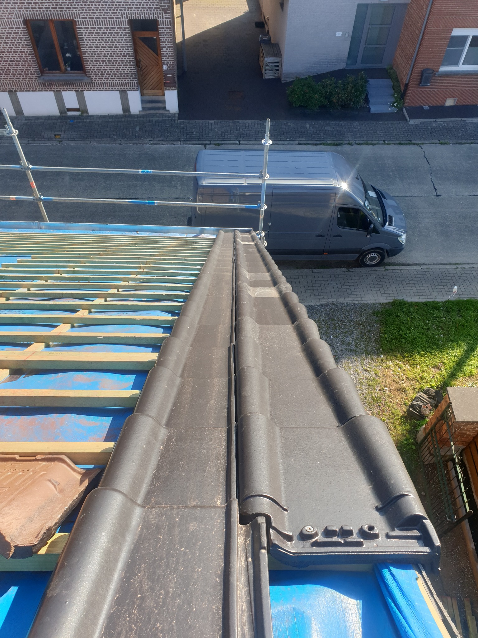 Widok z góry na fragment dachu w trakcie krycia, z widoczną łatą, membraną i częściowo ułożoną, ciemną dachówką ceramiczną, na tle ulicy z zaparkowanym szarym busem i fragmentami budynków.