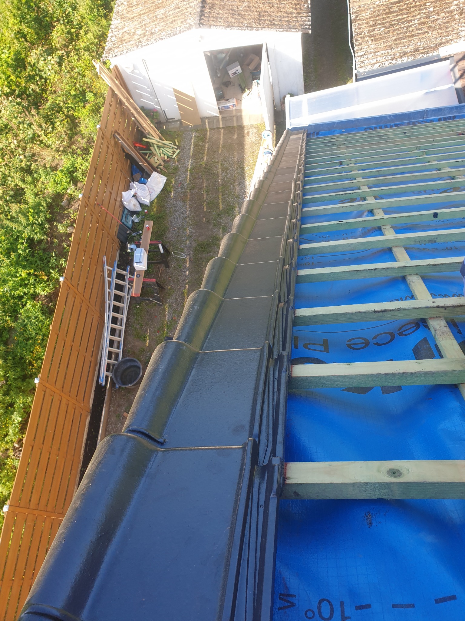 Układanie czarnej dachówki ceramicznej na łatach pokrytych niebieską membraną dachową, widok z góry na ogrodzenie z desek i budynek gospodarczy w tle.