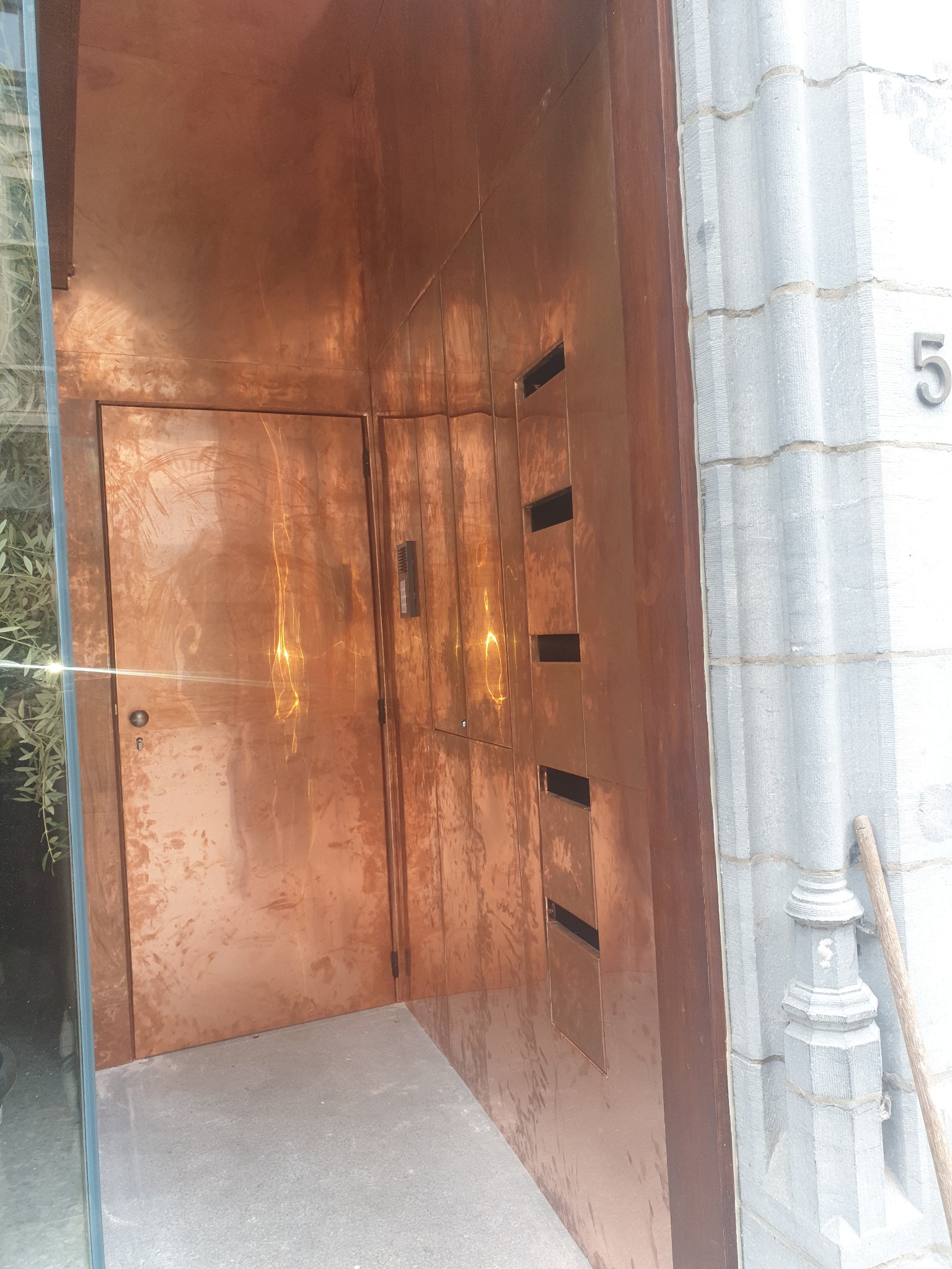 Miedziane wykończenie elewacji z drzwiami i skrzynkami pocztowymi, szara kamienna ściana z numerem 5.