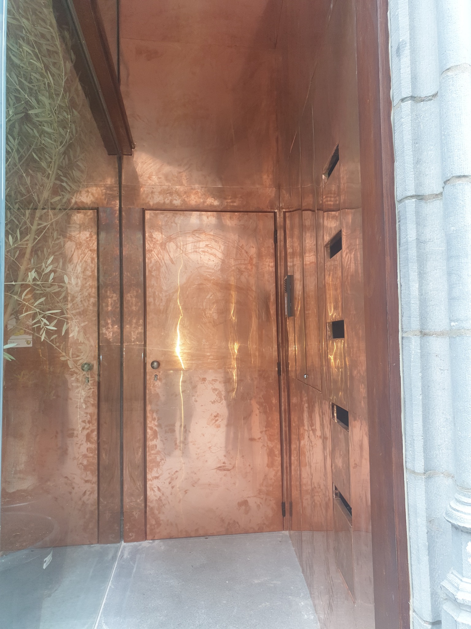 Miedziane drzwi wejściowe z wbudowanymi skrzynkami na listy, umieszczone w kamienicy z elementami dekoracyjnymi. Widoczne refleksy świetlne na metalowej powierzchni.