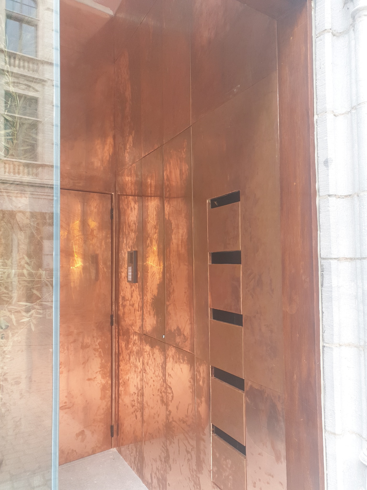 Miedziane wykończenie elewacji budynku z drzwiami i panelem z otworami wentylacyjnymi, widok z bliska z odbiciem budynku w szklanej powierzchni.