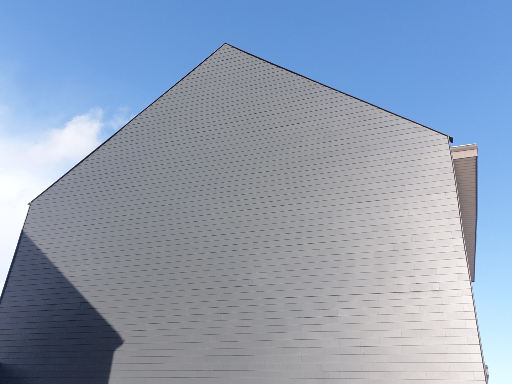 Szara, nowoczesna elewacja budynku z widoczną fakturą poziomych paneli, kontrastująca z błękitnym niebem i delikatnymi chmurami.