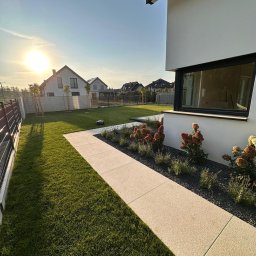 Dan-Bruk Woźnica Daniel - Nowoczesny ogród z trawnikiem, ścieżką z jasnych płyt i rabatami z ozdobnymi krzewami. W tle domy jednorodzinne i zachodzące słońce.