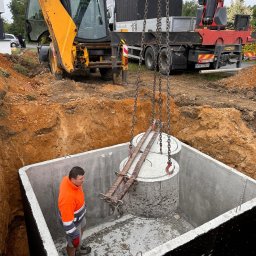 Dan-Bruk Woźnica Daniel - Montaż betonowego zbiornika w wykopie za pomocą dźwigu. Pracownik w odblaskowej kurtce nadzoruje proces. W tle koparka i ciężarówka.