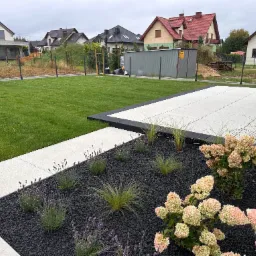 Nowoczesny ogród z trawnikiem, białymi płytami tarasowymi i rabatą z czarnym grysem, lawendą i hortensjami. W tle zabudowa jednorodzinna.