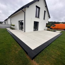 Dan-Bruk Woźnica Daniel - Nowoczesny taras z jasnych płyt, obramowany czarną kostką, przylegający do białego domu z czarną stolarką. Pomarańczowy samochód w tle. Świeża trawa.