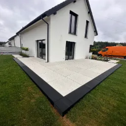 Nowoczesny taras z jasnych płyt, obramowany czarną kostką, przylegający do białego domu z czarną stolarką. Pomarańczowy samochód w tle. Świeża trawa.