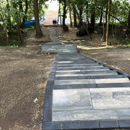 Dan-Bruk Woźnica Daniel - Schody z kostki brukowej prowadzące do drewnianego pomostu nad jeziorem, otoczone drzewami. Widok z góry na dół, z wyraźną teksturą kostki i otaczającej ziemi.