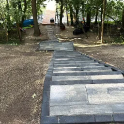 Schody z kostki brukowej prowadzące do drewnianego pomostu nad jeziorem, otoczone drzewami. Widok z góry na dół, z wyraźną teksturą kostki i otaczającej ziemi.