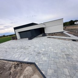 Dan-Bruk Woźnica Daniel - Nowoczesny podjazd z szarej kostki brukowej przed minimalistycznym domem z płaskim dachem. Widoczne obrzeża i przygotowany teren pod dalsze prace.