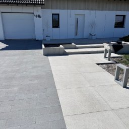 Dan-Bruk Woźnica Daniel - Nowoczesny podjazd z kostki brukowej w dwóch odcieniach szarości, prowadzący do białego domu z garażem i schodami. Minimalistyczne donice i oświetlenie.