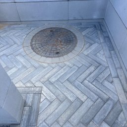 Dan-Bruk Woźnica Daniel - Szara kostka betonowa ułożona we wzór jodełki, z okrągłą studzienką w centrum. Widok z góry, podkreślający precyzję ułożenia i estetykę nawierzchni.