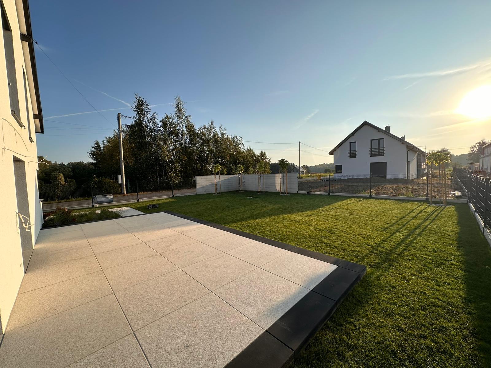 Jasny taras z płyt w ogrodzie, otoczony zielonym trawnikiem i nowoczesnym ogrodzeniem. W tle widoczny dom jednorodzinny w promieniach zachodzącego słońca.