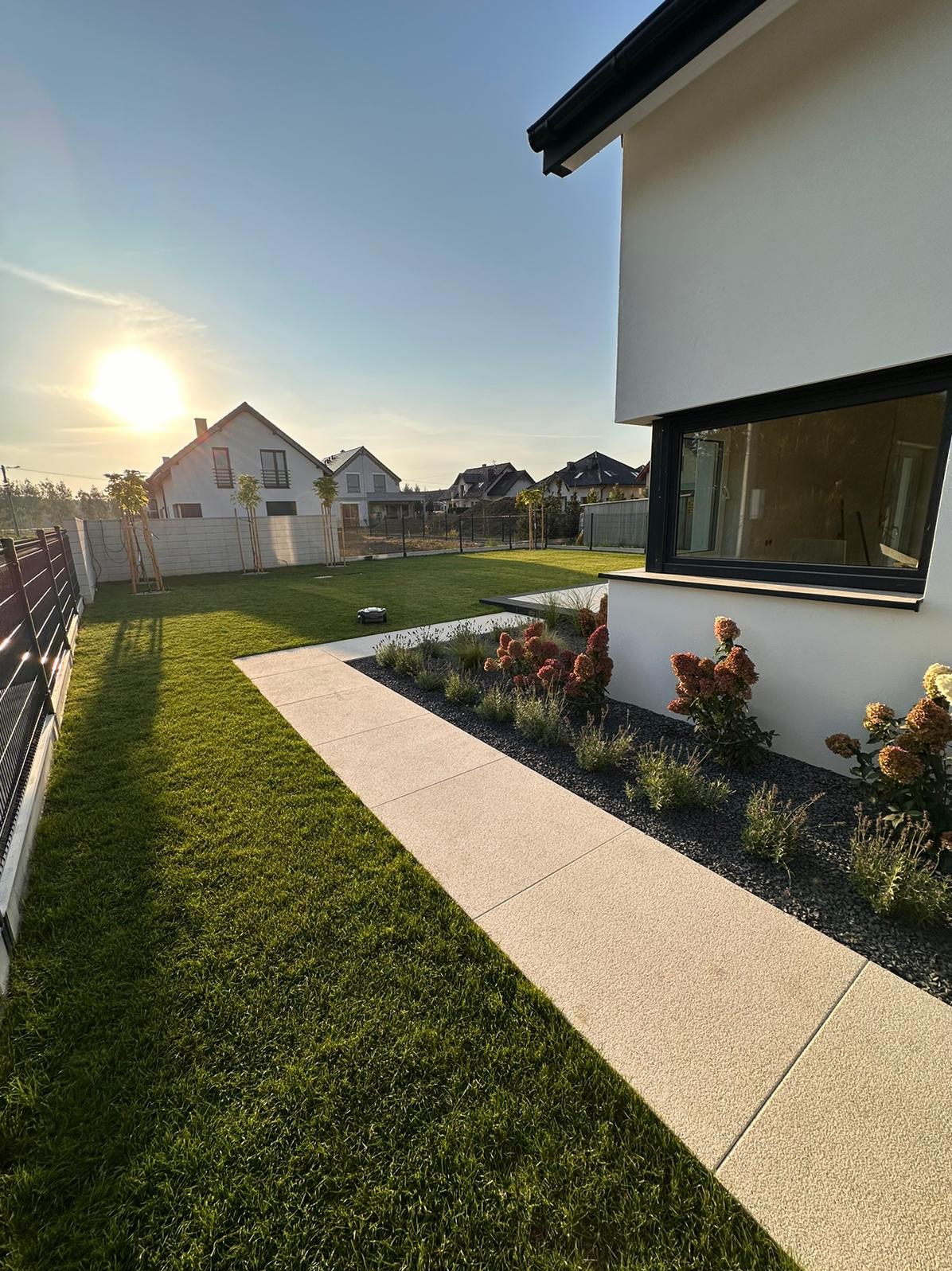 Nowoczesny ogród z trawnikiem, ścieżką z jasnych płyt i rabatami z ozdobnymi krzewami. W tle domy jednorodzinne i zachodzące słońce.