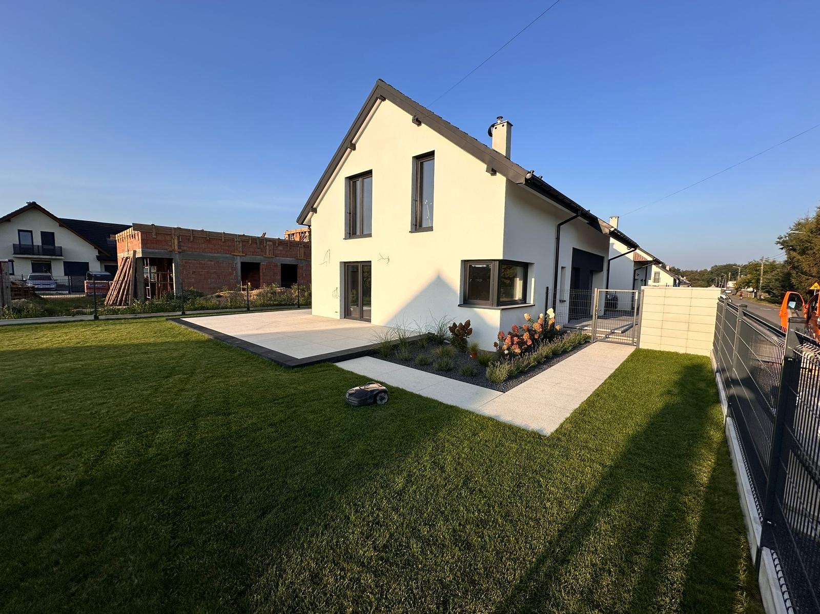 Nowoczesny dom z tarasem wyłożonym jasnymi płytami, zadbanym trawnikiem i robotem koszącym. Ścieżka z jasnego kamienia prowadzi do furtki. Widok na budowę w tle.