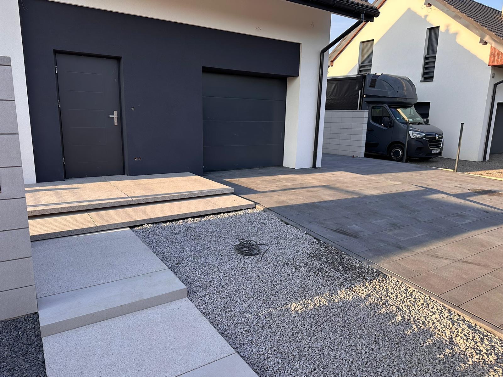 Nowoczesny podjazd z kostki brukowej w odcieniach szarości, schody wejściowe z jasnych płyt. Minimalistyczny design, widoczny fragment domu i zaparkowany samochód.