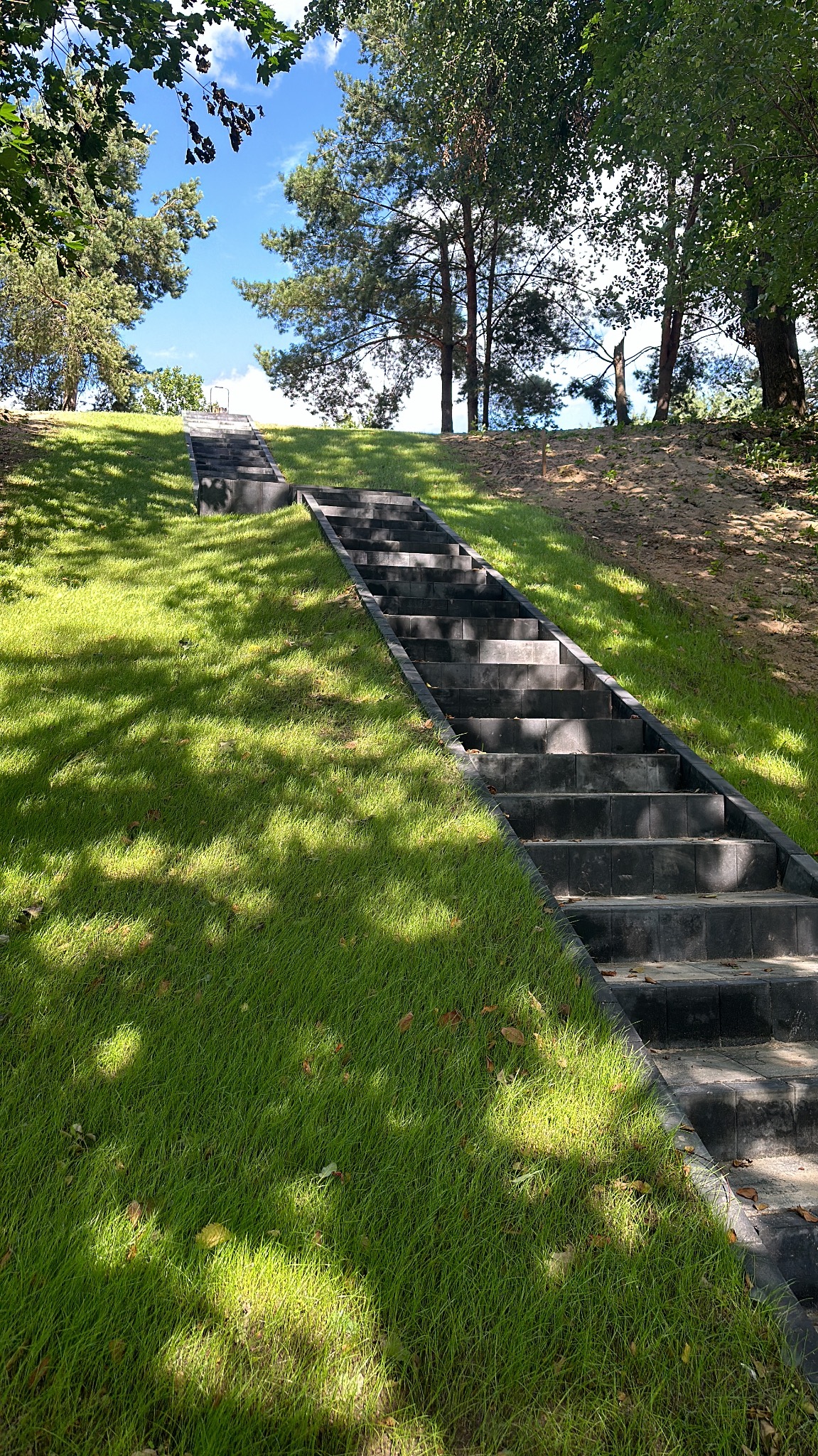 Schody z kostki brukowej prowadzące na wzgórze, otoczone zielonym trawnikiem i drzewami w słoneczny dzień. Widok z dołu, podkreślający perspektywę.