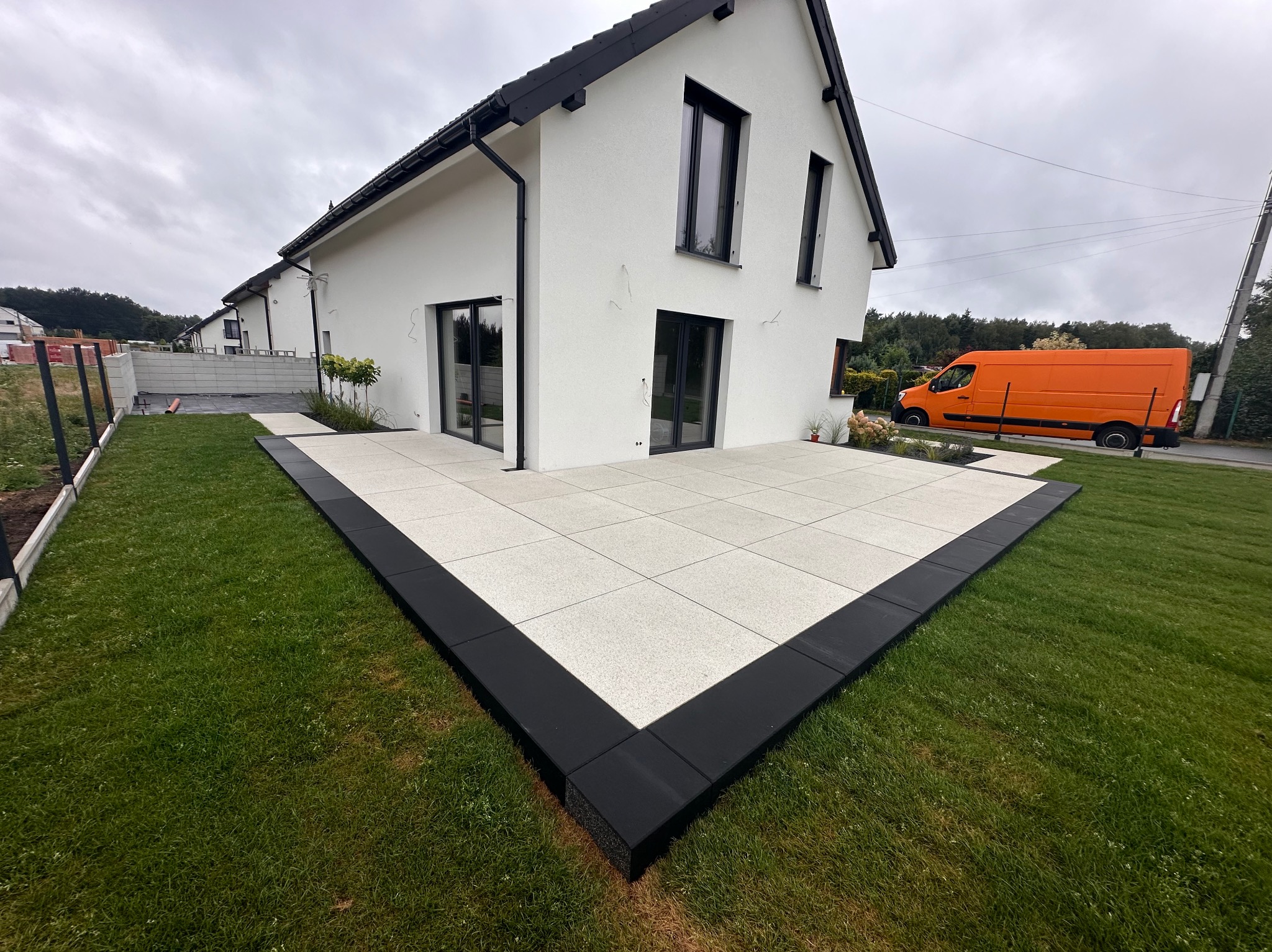 Nowoczesny taras z jasnych płyt, obramowany czarną kostką, przylegający do białego domu z czarną stolarką. Pomarańczowy samochód w tle. Świeża trawa.