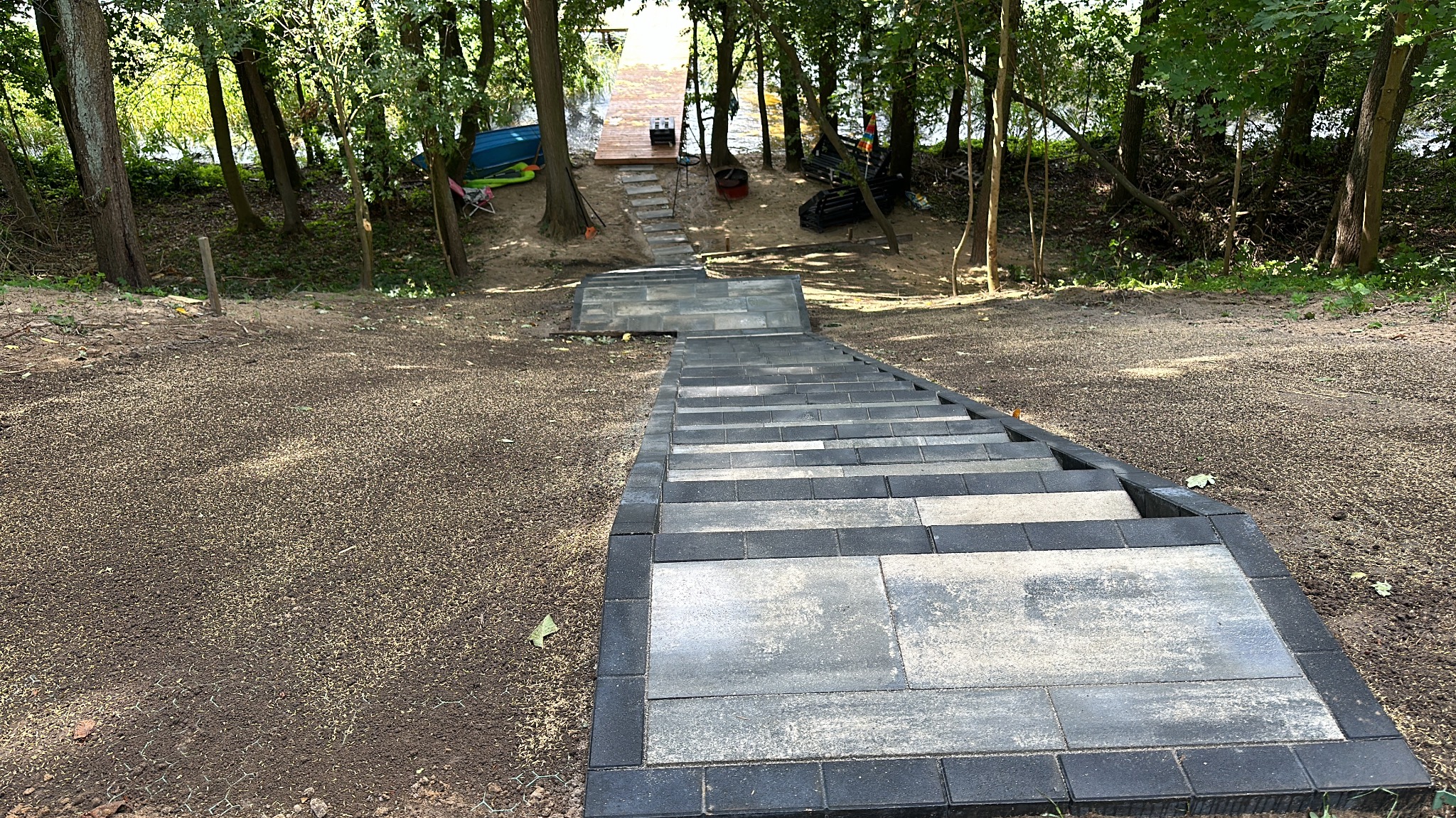 Schody z kostki brukowej prowadzące do drewnianego pomostu nad jeziorem, otoczone drzewami. Widok z góry na dół, z wyraźną teksturą kostki i otaczającej ziemi.