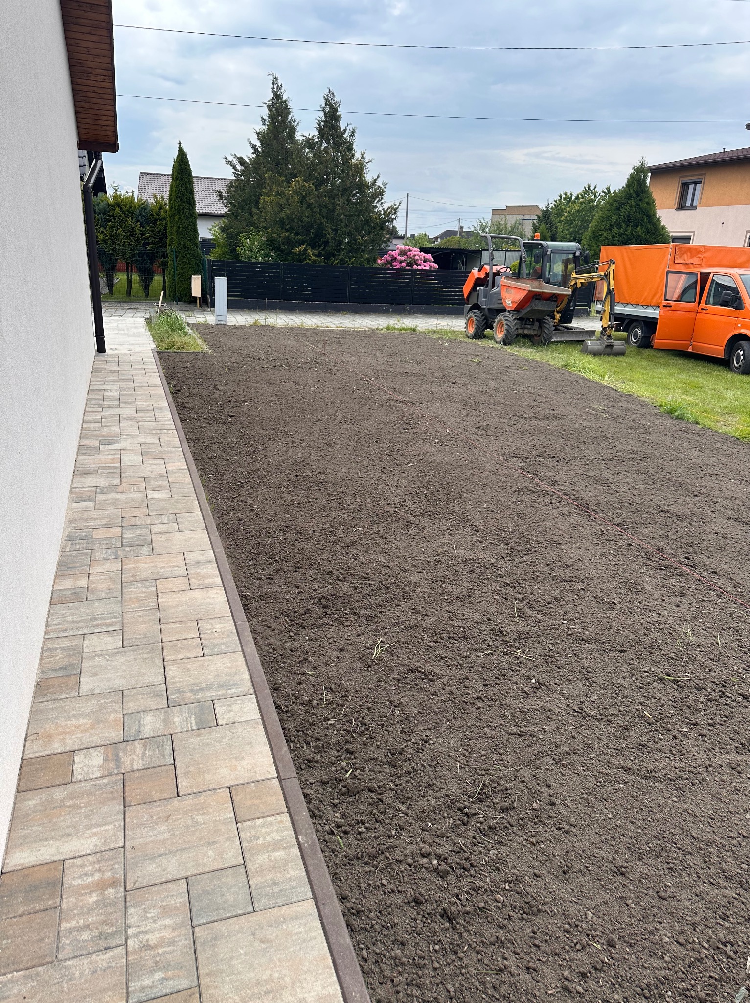 Przygotowany teren pod trawnik z mini koparką i ułożony chodnik z kostki w odcieniach beżu i szarości. Na drugim planie budynki mieszkalne i drzewa.