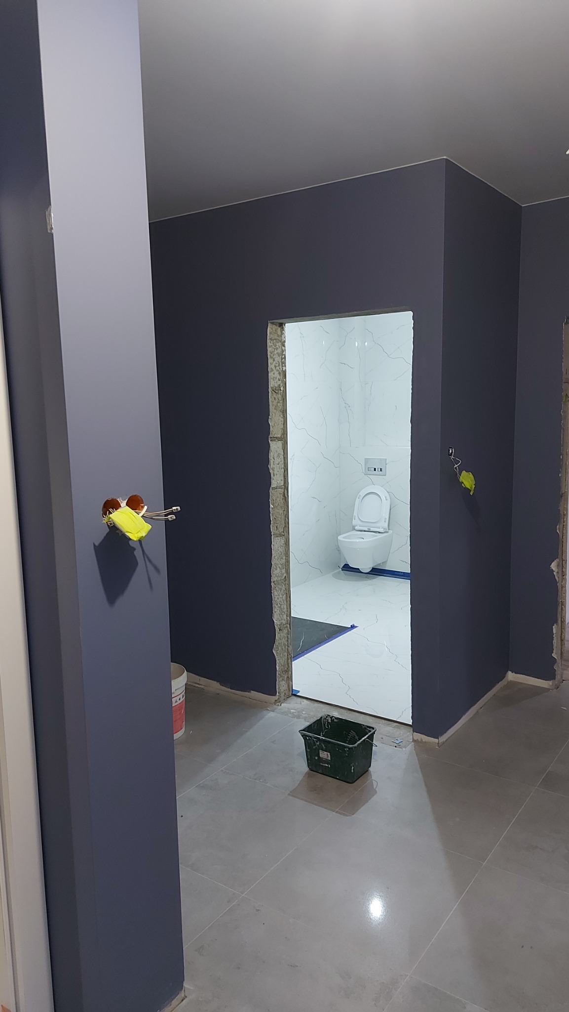 Wnętrze w trakcie remontu: świeżo pomalowane na ciemny granat ściany, widoczne puszki elektryczne z zabezpieczeniami, wiadro z farbą i otwarte drzwi do wykończonej toalety z białą armaturą.