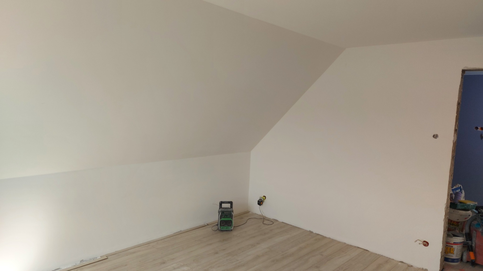 Pomalowane na biało poddasze ze skosami, widoczny grzejnik elektryczny i niezabezpieczone gniazdko elektryczne, w tle otwór drzwiowy z niebieską ścianą.