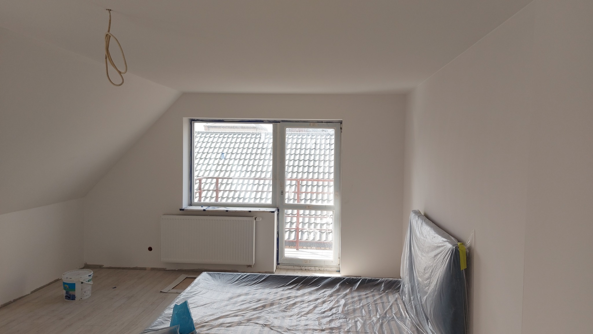 Pokój na poddaszu po świeżym pomalowaniu ścian na biało, z zabezpieczonym folią łóżkiem i grzejnikiem, widoczne zwisające kable oświetleniowe i wiadro z farbą.