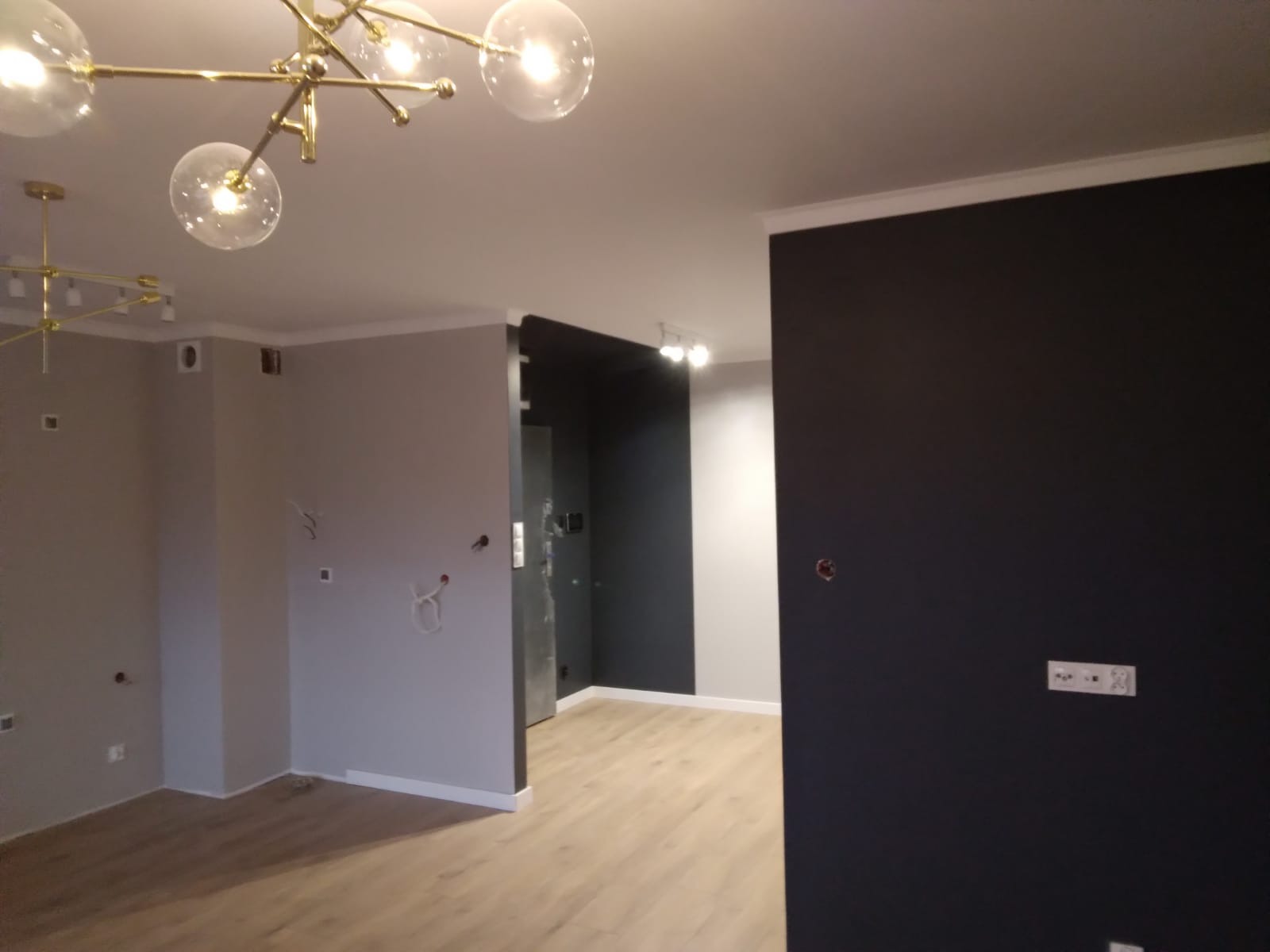 Wnętrze mieszkania w trakcie malowania, widoczne szare ściany z wystającymi kablami, jedna ściana pomalowana na ciemny grafit, drewniana podłoga i nowoczesna lampa sufitowa.