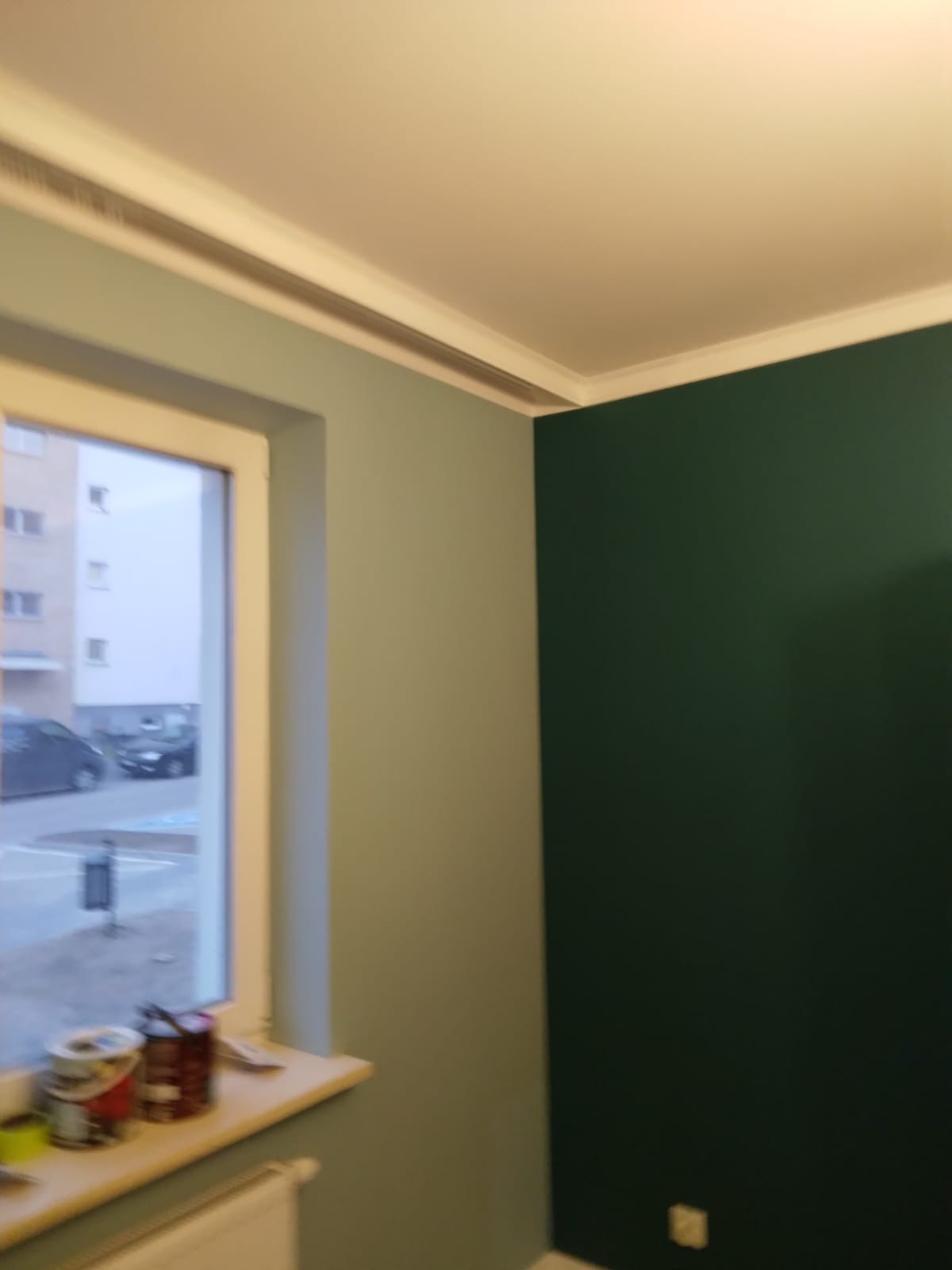 Pomieszczenie po malowaniu: ściany w odcieniach zieleni, jaśniejsza przy oknie, ciemniejsza w rogu, widoczne puszki z farbą na parapecie.