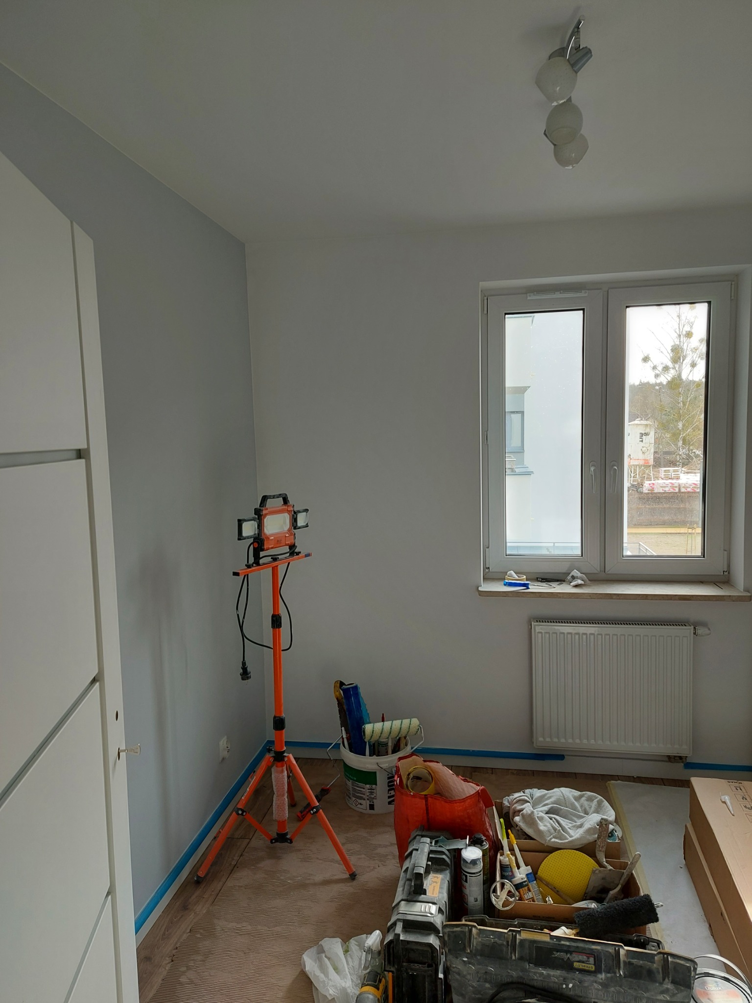 Pokój w trakcie odświeżania: ściany pomalowane na szaro, widoczna lampa budowlana na trójnogu, narzędzia malarskie i wiadro z farbą, zabezpieczona taśmą podłoga, grzejnik pod oknem.
