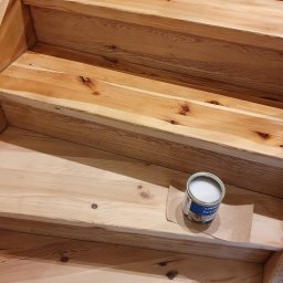 Gduli-Bud - Drewniane schody po renowacji, widoczna puszka z lakierem do drewna na papierze ochronnym, jasne drewno z widocznymi słojami.