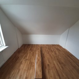 Gduli-Bud - Poddasze po remoncie z jasnymi ścianami i nową, drewnianą podłogą z widocznym fragmentem nieułożonych paneli oraz grzejnikiem i oknem w tle.