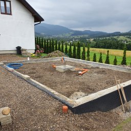Gduli-Bud - Przygotowany fundament pod budowę domu z widocznymi instalacjami, izolacją przeciwwilgociową, ziemią i gruzem, na tle górskiego krajobrazu z tujami.