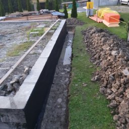 Gduli-Bud - Betonowa podmurówka fundamentowa z hydroizolacją pionową w wykopie, widoczne materiały budowlane, ziemia i trawa na działce.