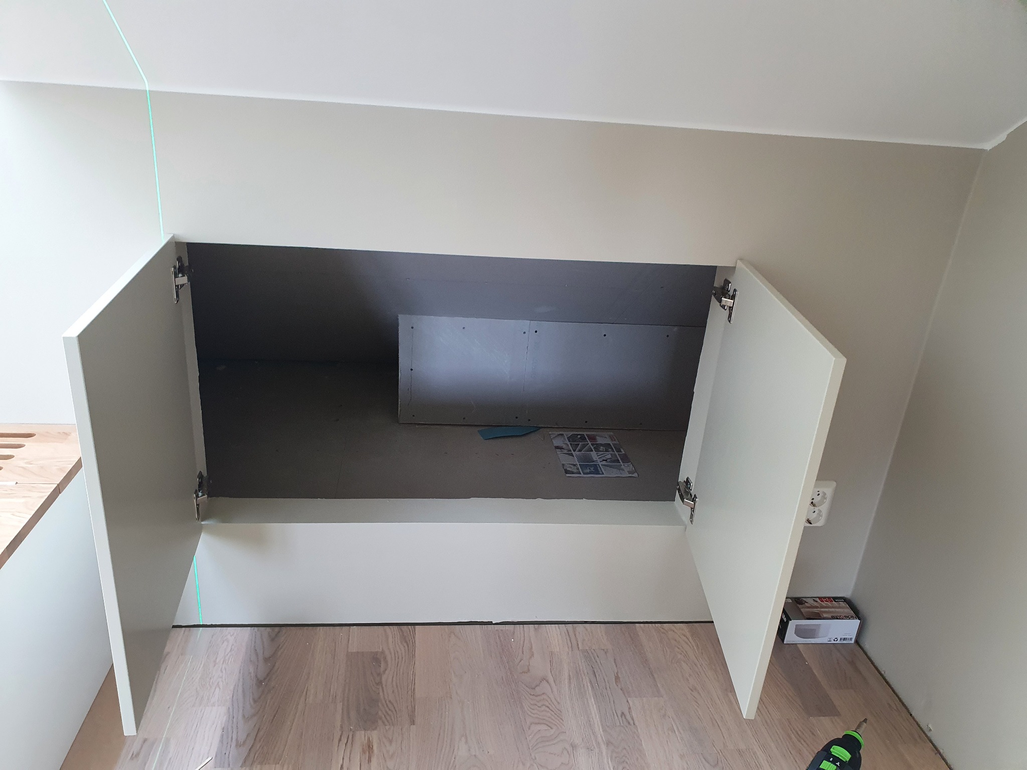 Wnęka w ścianie zabudowana szafką z otwartymi drzwiczkami, widoczna zielona linia lasera, podłoga wyłożona panelami, w środku wnęki płyta karton gips.