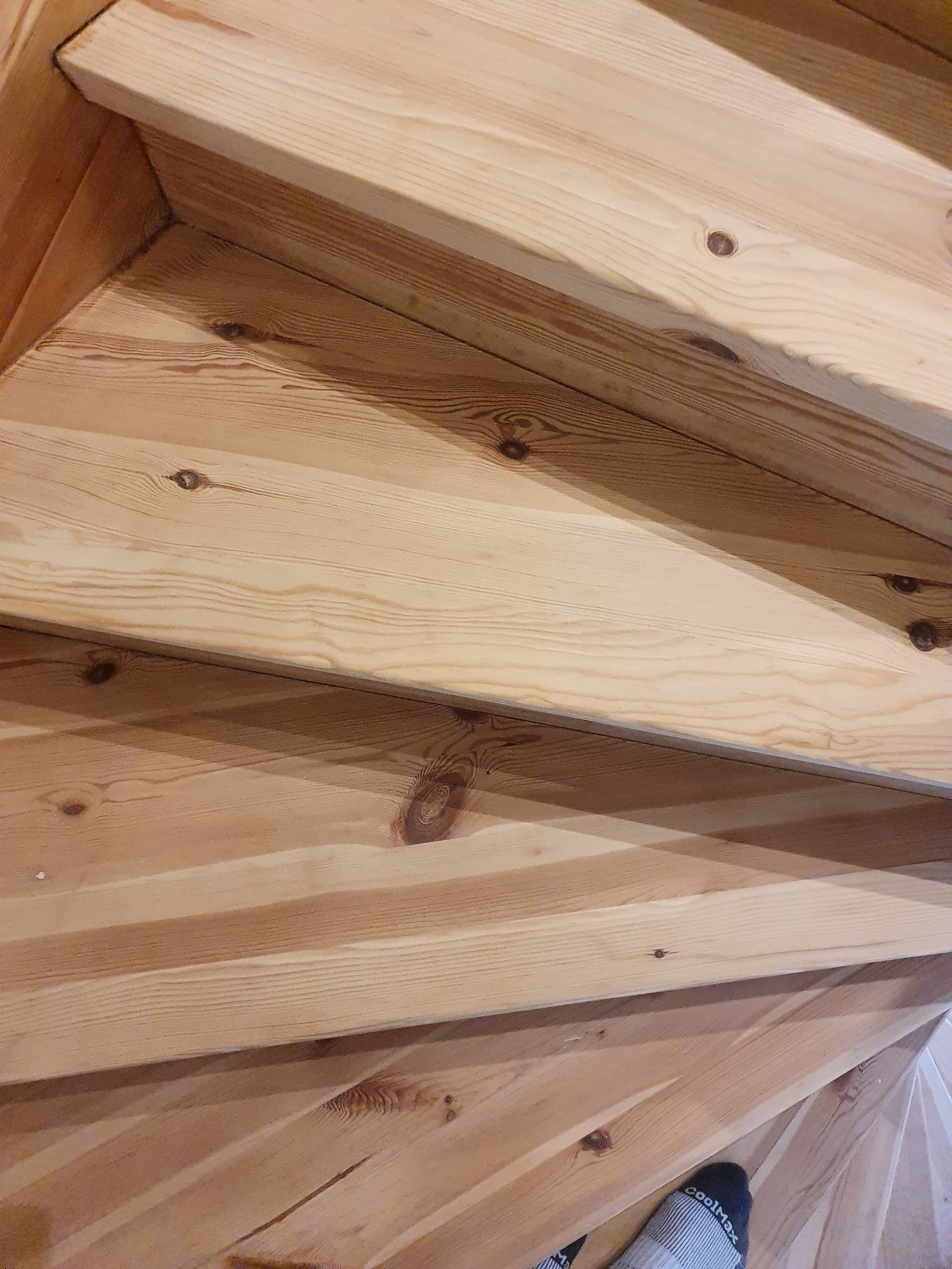 Fragment drewnianych schodów kręconych z widocznymi słojami i sękami, ujęcie z góry z fragmentem stopy w skarpetce na dolnym planie.