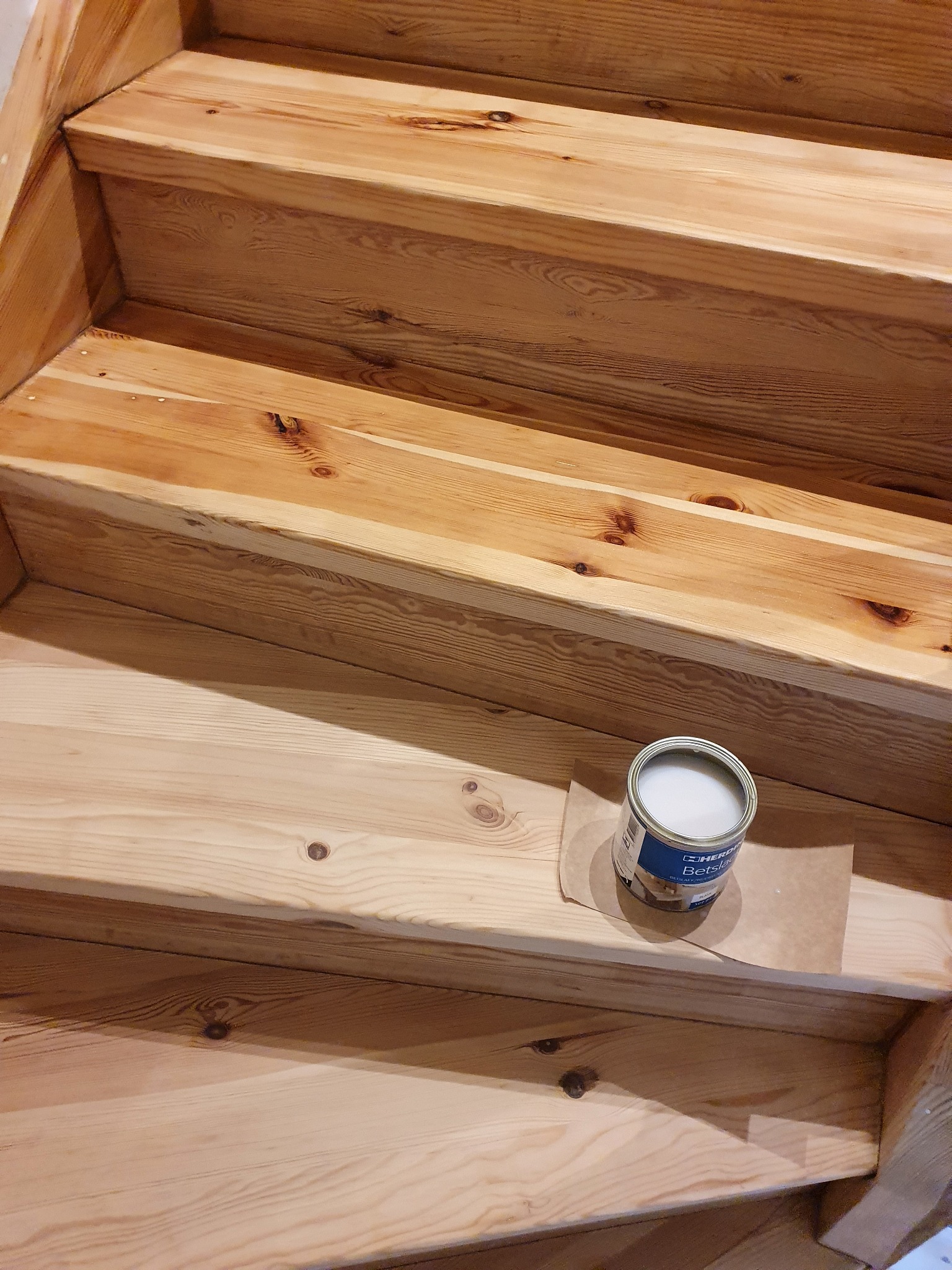 Drewniane schody po renowacji, widoczna puszka z lakierem do drewna na papierze ochronnym, jasne drewno z widocznymi słojami.