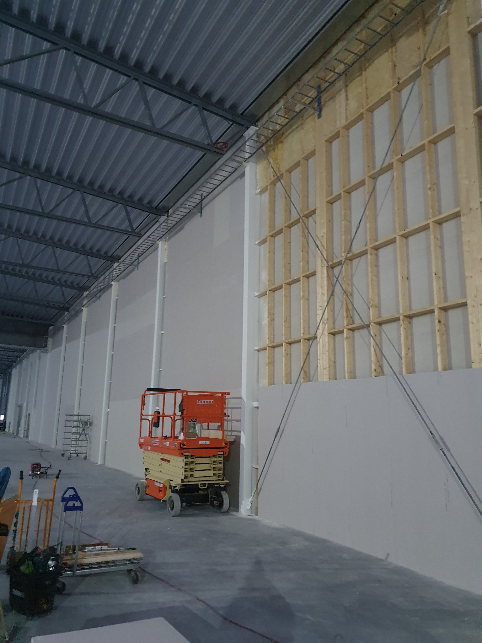 Wnętrze hali produkcyjnej w trakcie montażu ścian z płyt gipsowo-kartonowych na drewnianej konstrukcji szkieletowej, widoczny podnośnik nożycowy i stalowa konstrukcja dachu.