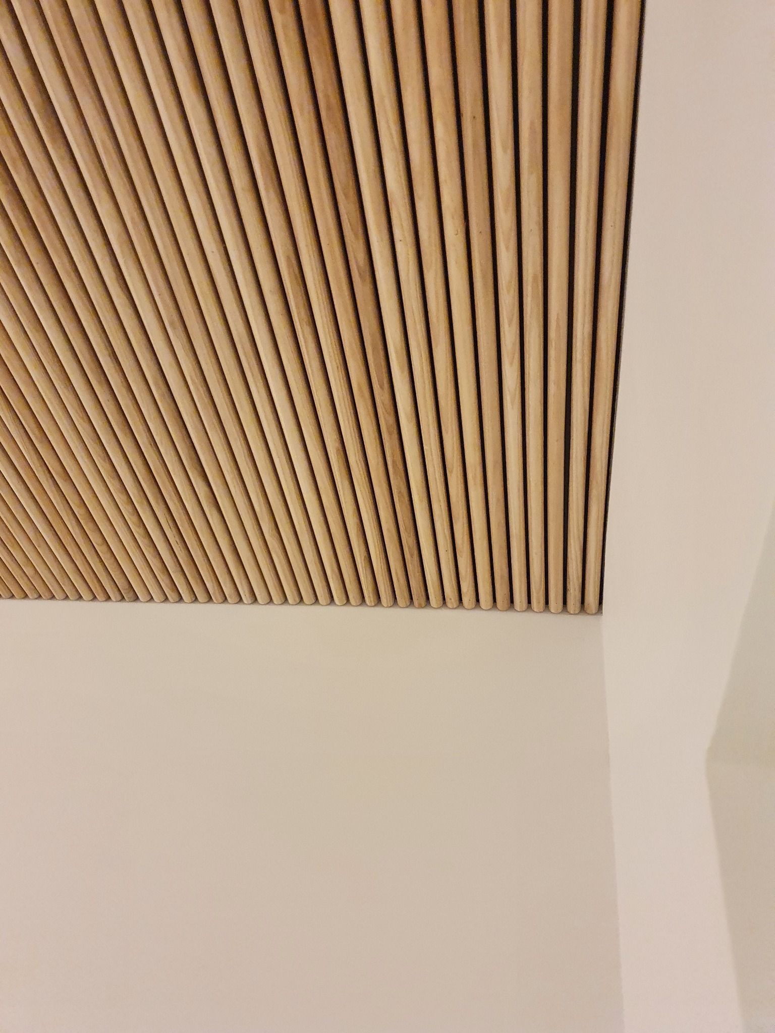 Drewniane listwy dekoracyjne na suficie, ułożone równolegle pod kątem, przylegające do gładkiej, beżowej ściany.