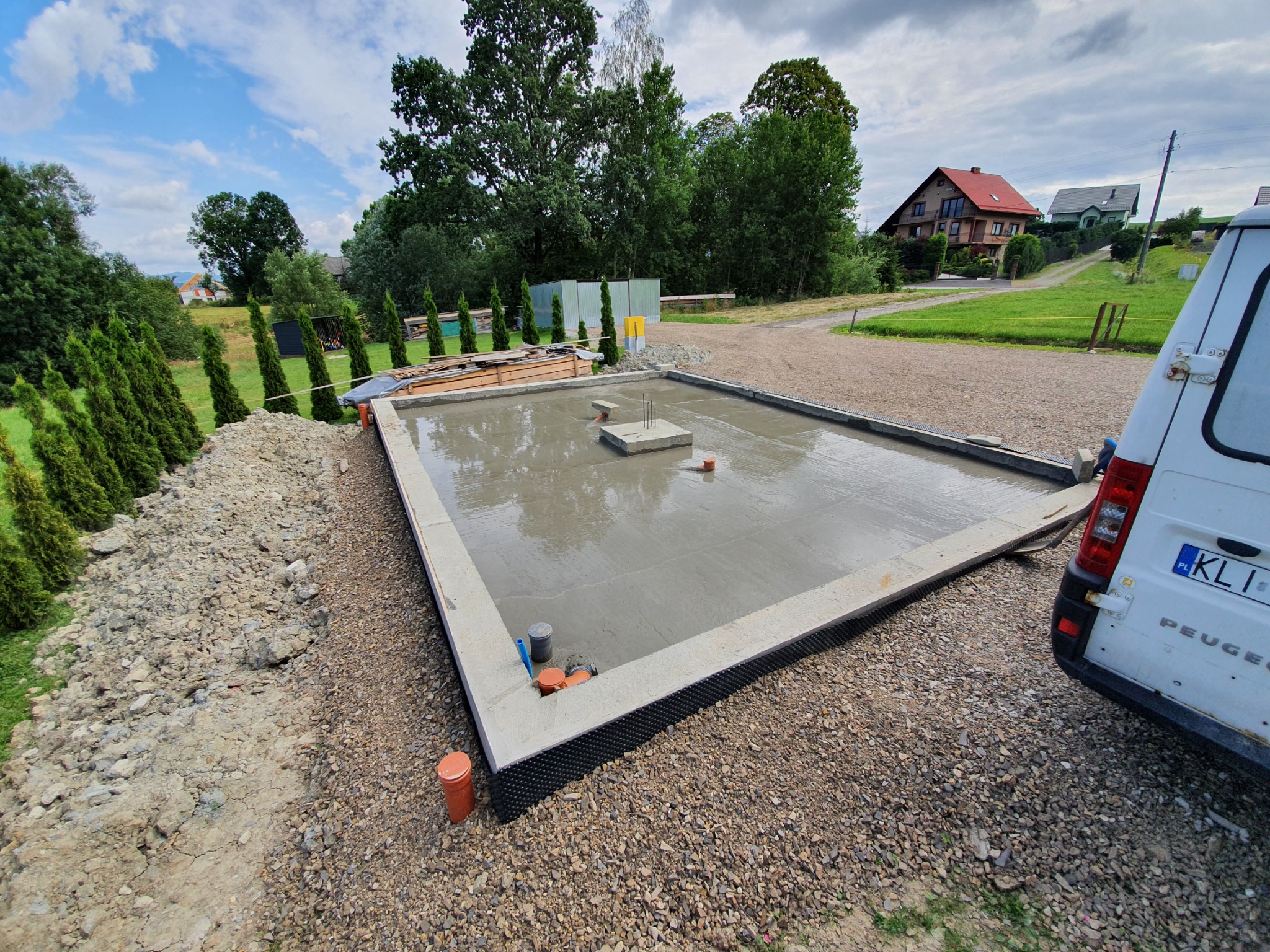 Świeżo wylana płyta fundamentowa z wystającymi zbrojeniami, zabezpieczona folią kubełkową, widok z boku, zaparkowany samochód dostawczy w tle.