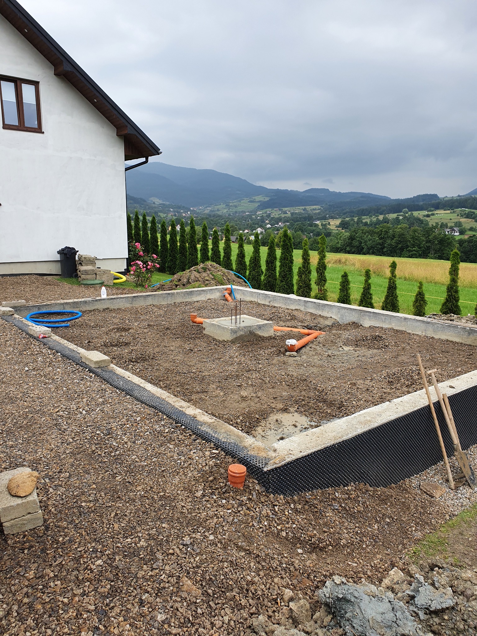 Przygotowany fundament pod budowę domu z widocznymi instalacjami, izolacją przeciwwilgociową, ziemią i gruzem, na tle górskiego krajobrazu z tujami.